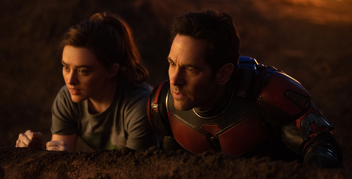 Cassie Lang, played by Kathryn Newton, and Scott Lang, played by Paul Rudd, are crouched over the edge of a wall. Newton is wearing a gray T-shirt and has her hair in a ponytail. Rudd is wearing a red, black, and silver Ant-Man suit.