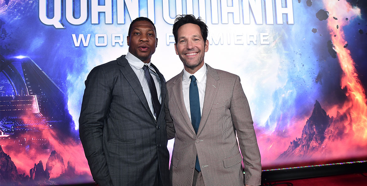 Jonathan Majors and Paul Rudd pose together in front of a large Ant-Man and The Wasp: Quantumania backdrop. Majors has on a dark gray suit with matching tie. Rudd is wearing a brown and white checkered suit with a navy-blue tie. Rudd is smiling at the camera while Majors has a slight pout on his face.