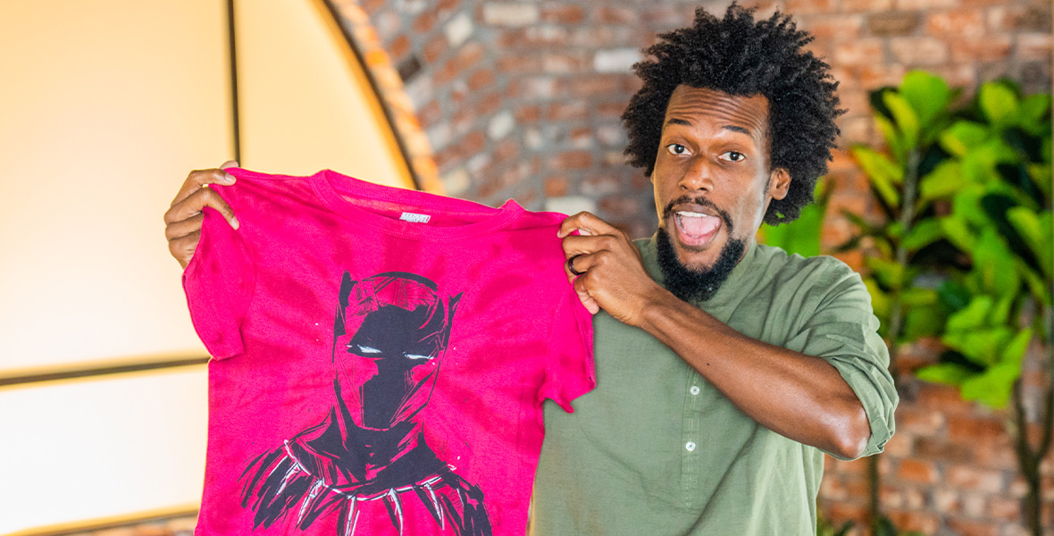 Nikkolas Smith, with an excited expression, holds up a red t-shirt with his art of Marvel’s Black Panther on it.