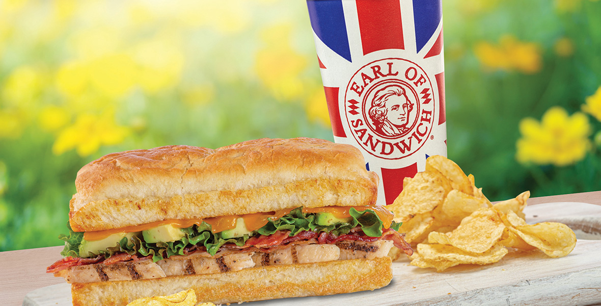 In a promotional image for Earl of Sandwich, a chicken sandwich on toasted bread—with avocado, bacon, lettuce, and tomato—is seen on a wooden serving tray to the left, surrounded by potato chips. To the right and slightly behind the tray is an Earl of Sandwich drink cup, which features the restaurant’s red, white, and blue logo, depicting a man with the words “Earl of Sandwich” around his face. Behind the sandwich and drink is a field of blurred yellow flowers.