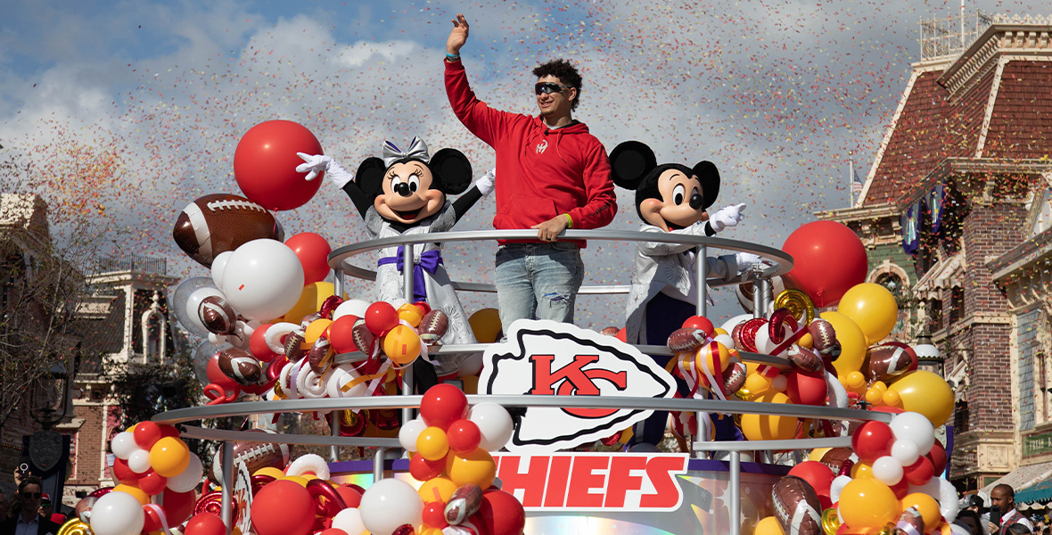 A photo from the Main Street, U.S.A. parade celebrating MVP and Kansas City Chiefs quarterback Patrick Mahomes at Disneyland Park. Mahomes is wearing a red hoodie and is standing on a float and waving to the crowd; Minnie Mouse (on left) and Mickey Mouse (on right) are standing on either side of him, wearing their Disney100 outfits. There are red, yellow, white, and football-shaped balloons adorning the float, as well as a Chiefs logo. Red and yellow paper confetti can be seen floating in the air.