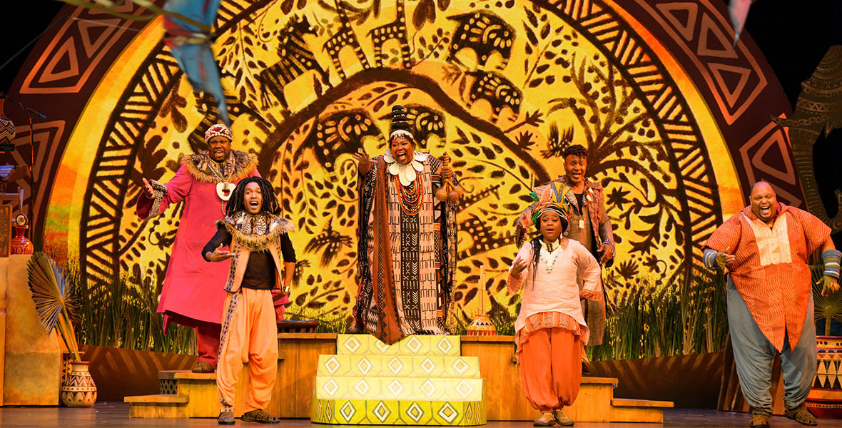 A photo of Tale of the Lion King, where six performers sing toward the audience. They wear traditional African-inspired clothing, with a brilliant yellow backdrop behind them. Silhouettes like giraffes, zebras, vines, and leaves adorn the backdrop, which shines in a half-circle like the rising sun.