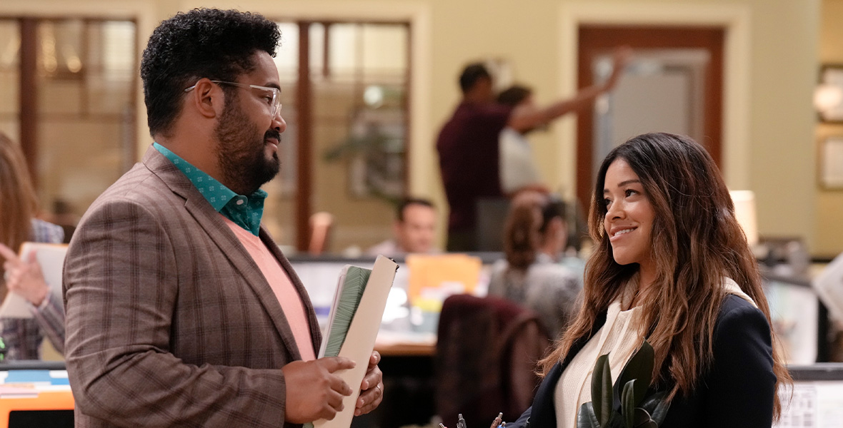 In a scene from the ABC series Not Dead Yet, actors Josh Banday as Dennis and Gina Rodriguez as Nell talk in a newspaper newsroom. Banday wears a teal button-down shirt, a pink sweater, and a brown suit jacket and holds file folders. Rodriguez wears a white blouse and a black suit and holds a coffee mug and potted plant.