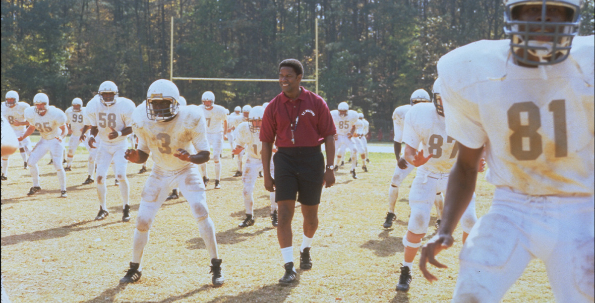 In a scene from the feature film Remember the Titans, actor Denzel Washington as head coach Herman Boone wears a maroon polo shirt, black shorts, and a whistle attached to a black lanyard around his neck. Washington is surrounded by football players wearing white jerseys and white pants.