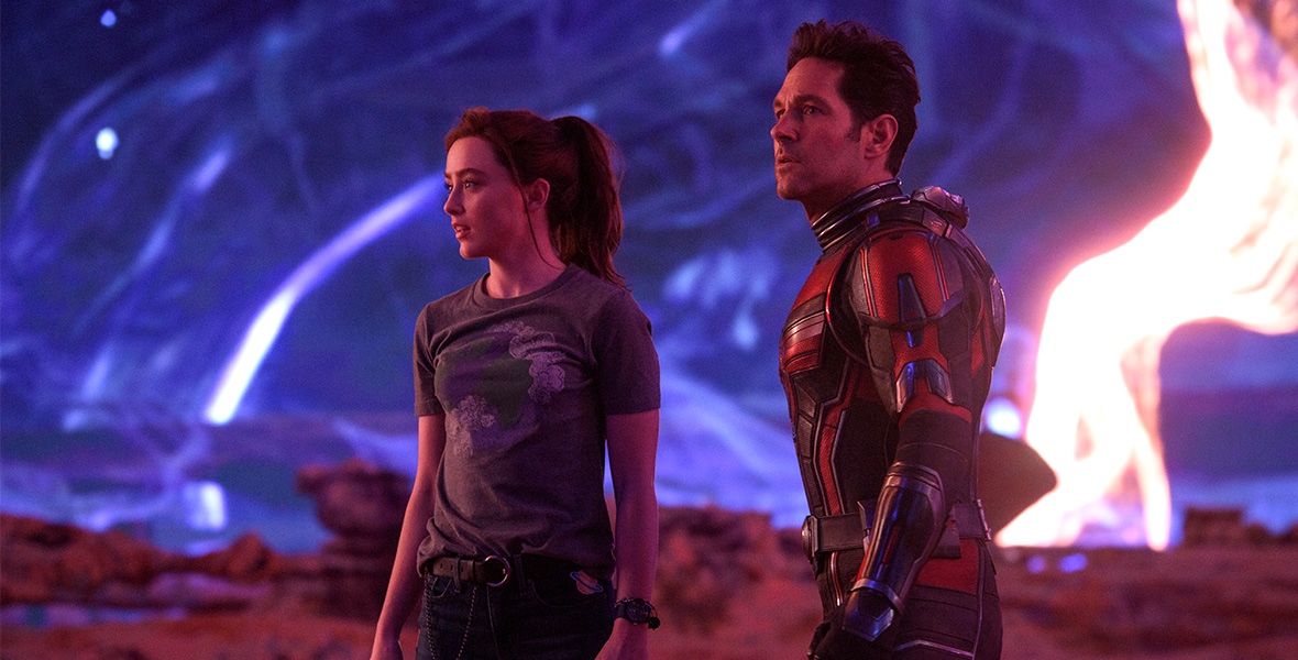 In a scene from Ant-Man and The Wasp: Quantumania, Cassie Lang (left) and Scott Lang/Ant-Man (right) stand and look to their right. Cassie has tucked a gray T-shirt into skinny jeans and accessorized with a belt. Her brown hair is pulled back into a high ponytail. Scott is wearing a black and red suit; his helmet is off. They are in the Quantum Realm, which includes red rocks, a purple sky, and electric lights in the sky.