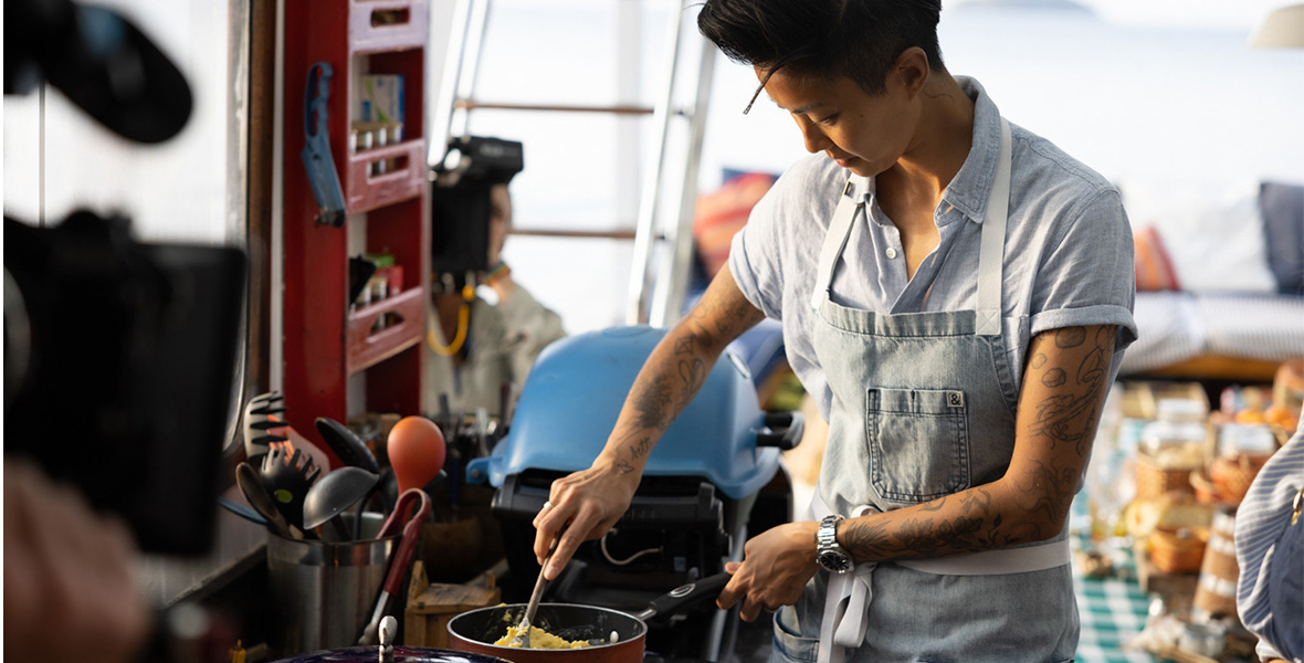 In a scene from Restaurants at the End of the World, Kristen Kish cooks eggs in an outdoor kitchen. She is using one hand to turn the eggs with a spatula and using the other hand to steady the pot. She is wearing a denim apron over a chambray shirt.