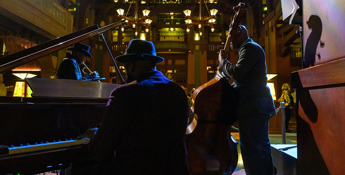 Under the high ceilings of the Great Hall of Disney’s Grand Californian Hotel & Spa, we see the backs of three performers. The one closest to the camera plays the piano, while another strums the cello and one blows the trumpet.