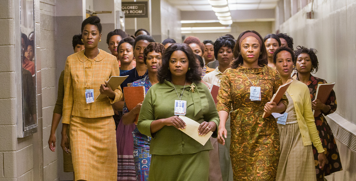 In a scene from Hidden Figures, actor Octavia Spencer as Dorothy Vaughan leads a group of Black women down a long hallway. She wears a green blouse and a matching green skirt and holds files in her hands.