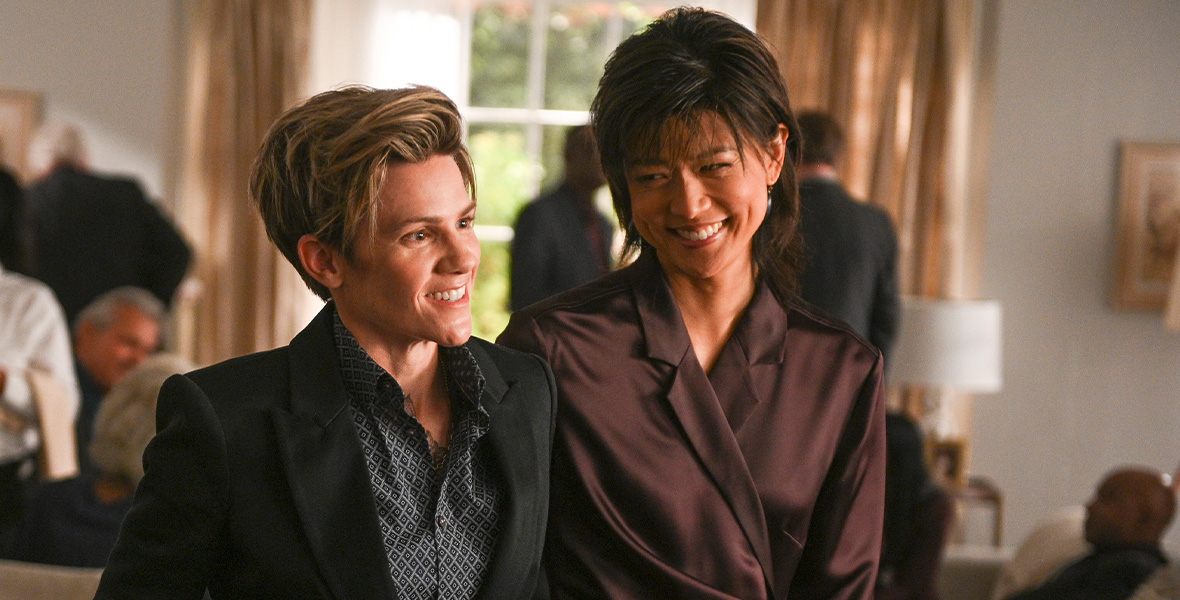 In a scene from the ABC series A Million Little Things, actors Cameron Esposito as Greta and Grace Park as Katherine stand side by side inside a home. Esposito wears a black suit and a black and white patterned button-down top. Park wears a brown silk wrap dress and smiles at Esposito.