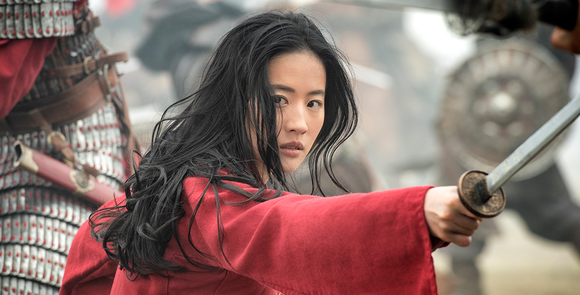 In a scene from Mulan, actor Yifei Liu as Mulan wears a red tunic and yields a large sword in her right hand in a battle field.