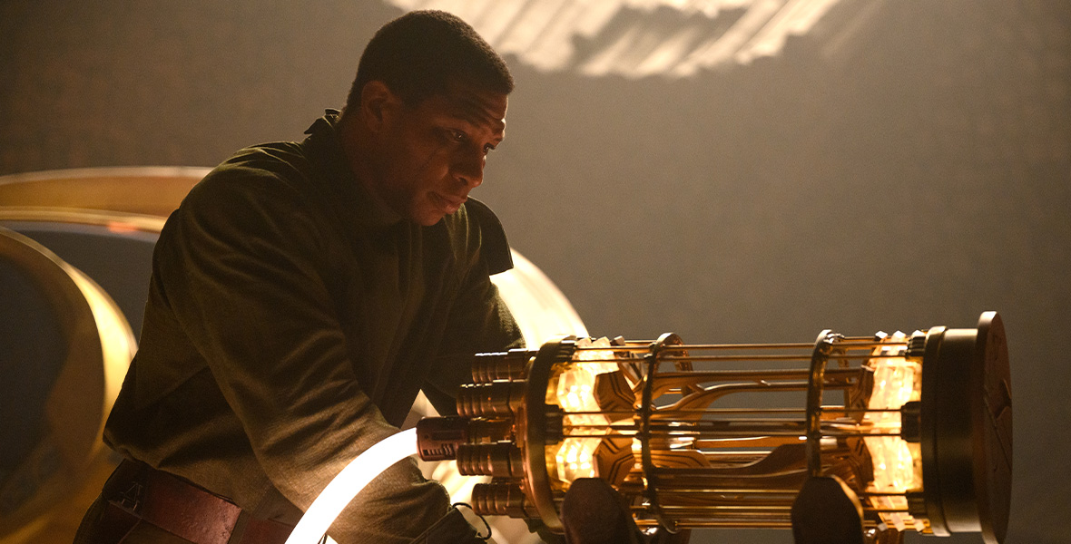 Kang the Conqueror leans with his hands against a gold metallic device, staring down in thought. He wears a green jacket and brown belt with close-cropped dark hair.