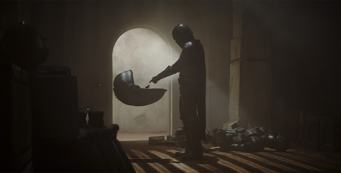 In an image from The Mandalorian, Mando is seen pointing towards The Child—later revealed to be Grogu—in his floating carriage; Grogu is pointing back. Both are backlit by the light streaming through a doorway behind them.