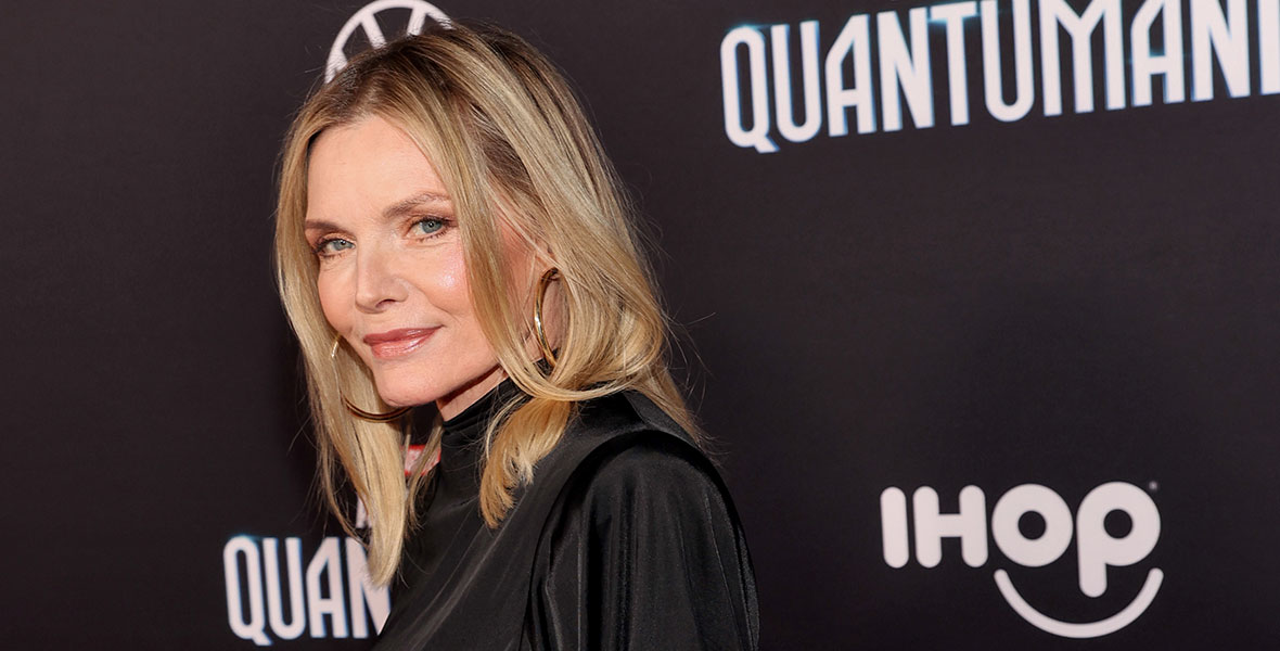 Michelle Pfeiffer stands in front of a black backdrop with a closed mouth smile on her face. She is wearing a draped black long sleeve dress and large gold hoop earrings. Behind her you can see the logo for Ant-Man and The Wasp: Quantumania.
