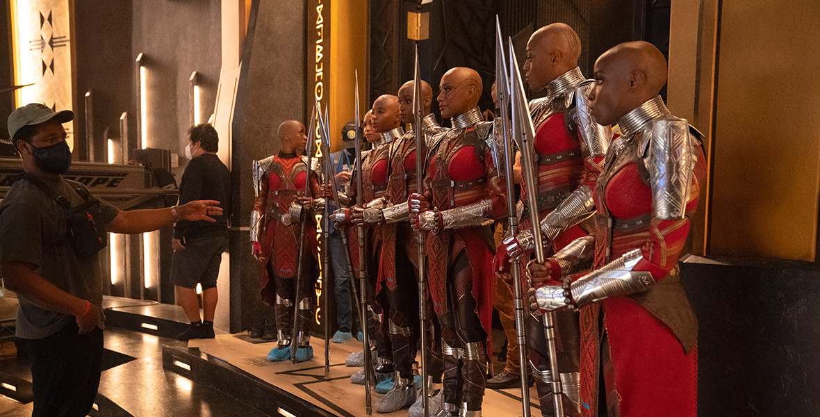 Director Ryan Coogler gestures to actors portraying the Dora Milaje, a group of warriors, before filming a scene for Marvel Studios’ Black Panther: Wakanda Forever. Coogler wears a tan baseball hat, an olive T-shirt, black pants, and a black face mask.