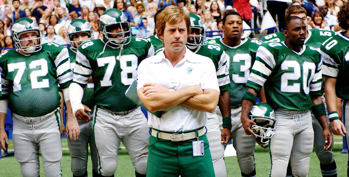 In a scene from the feature film Invincible, actor Greg Kinnear as NFL coach Dick Vermeil stands on the side with his arms folded across his chest. He wears a white polo shirt, a white belt, and green pants. Behind Kinnear are men portraying Philadelphia Eagles football players and a stadium crowd.