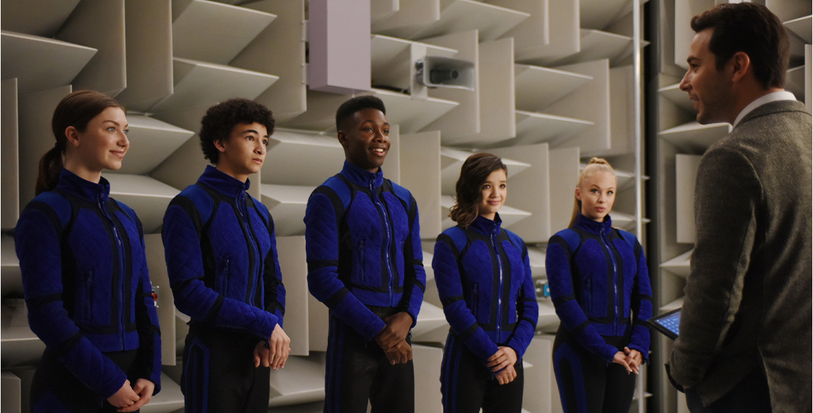 In a scene from Secret Society of Second-Born Royals, actors Isabella Blake-Thomas, Faly Rakotohavana, Niles Fitch, Peyton Elizabeth Lee, and Olivia Deeble, stand in a line opposite actor Skylar Astin and wear royal blue and black jackets and black pants with royal blue stripes down the side. Astin wears a light brown tweed jacket and a white button-down shirt.