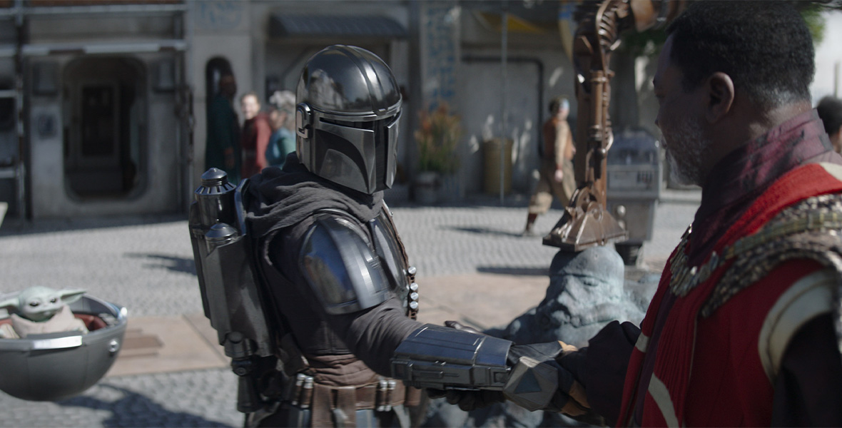 In a scene from an episode of Star Wars: The Mandalorian, actor Pedro Pascal as Mandalorian/Din Djarin shakes hands with actor Carl Weathers as Greef Karga. Pascal wears a metal helmet and metal suit of armor. Weathers wears a red tunic and a gold patterned cloak.