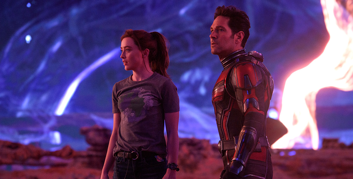 Cassie and Scott stand together as they look off into the distance. The purple sky and red-brown rocks of the Quantum Realm are behind them. Scott wears his red and black Ant-Man suit while Cassie sports a gray T-shirt and jeans. Her brown hair is pulled back into a ponytail.