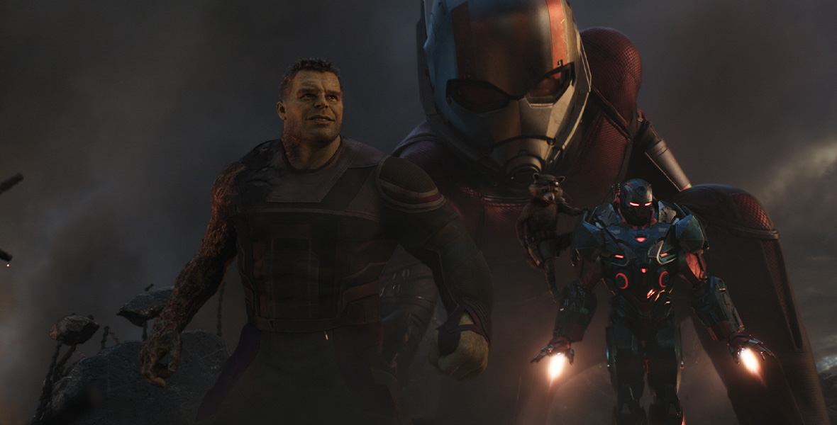 In a scene from Avengers: Endgame, Smart Hulk, Rocket Raccoon, and War Machine emerge from the rubble in the foreground. In the background, Ant-Man has turned into Giant-Man. Even while crouching, he towers over the other Super Heroes.