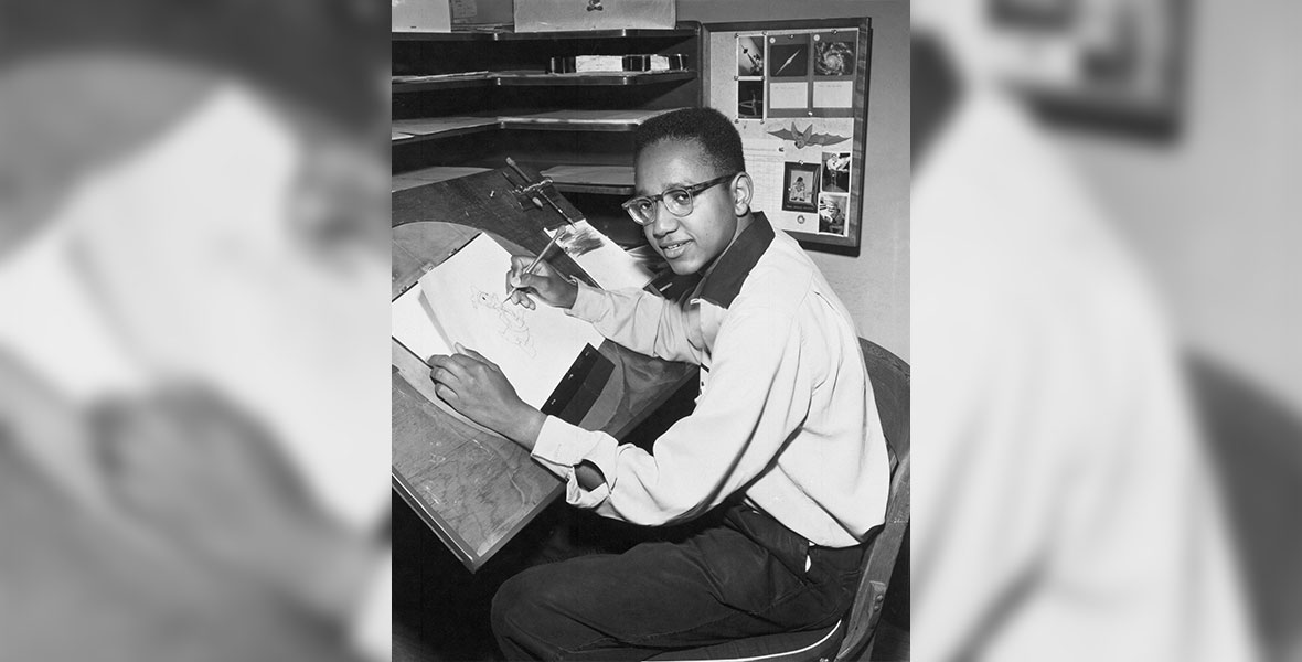 A black and white photo of Floyd Norman, who looks up from his half-done drawing of Donald Duck at the camera. He wears a white, long-sleeved shirt and glasses.