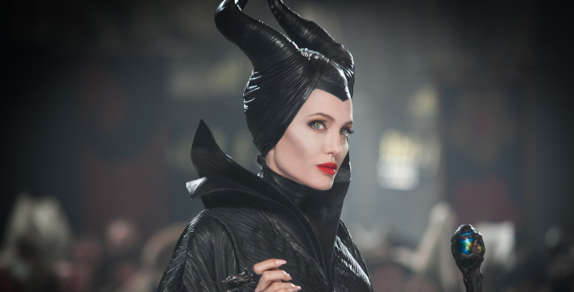 In a scene from Maleficent, actor Angelina Jolie as Maleficent looks over her right shoulder and holds a black staff in her left hand. She wears a black headpiece with two horns, a black cloak, and a black gown.