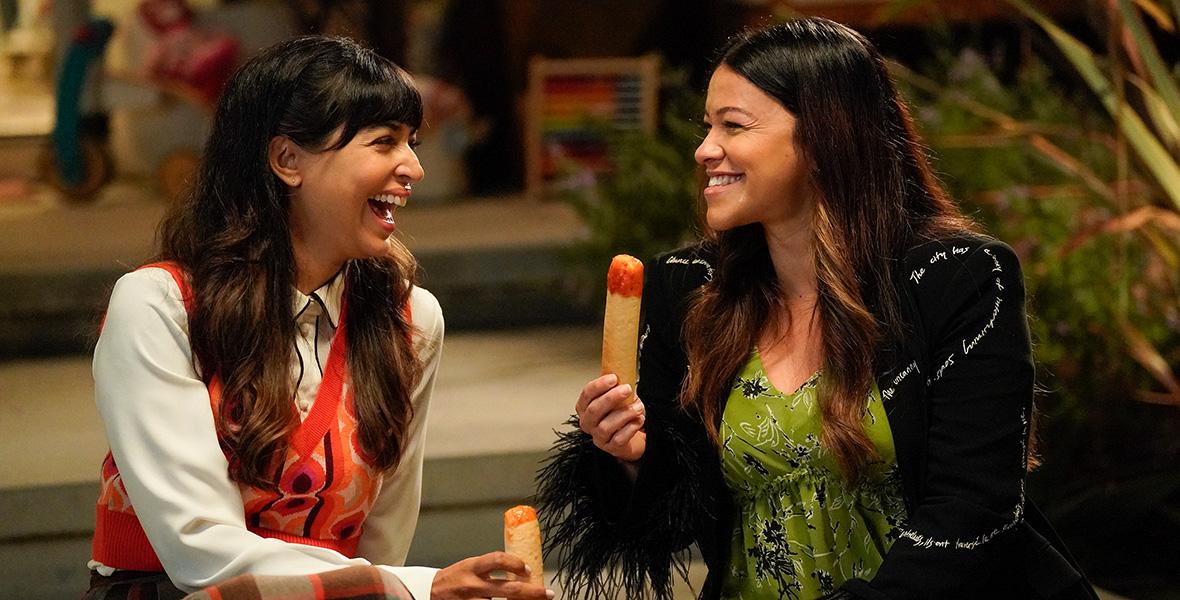 In a scene from an episode of Not Dead Yet, actors Hannah Simone as Sam and Gina Rodriguez as Nell sit on steps and eat breadsticks. Simone wears a white blouse, an orange patterned vest, and an orange and gray plaid skirt. Rodriguez wears a green floral dress and a black patterned sweater.