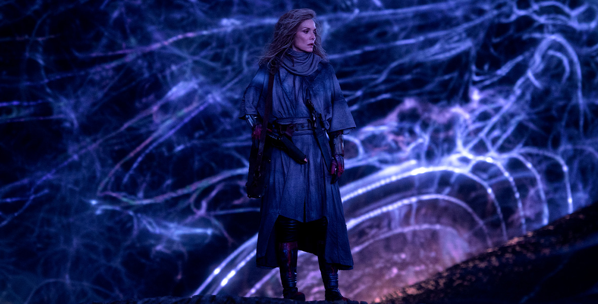 Janet stands on what looks to be a dark ridge, with the electric blue, black, and purple sky of the Quantum Realm behind her. She looks solemnly to her right. She’s clad in robes, gloves, and boots, and her blond hair brushes just past her shoulders.