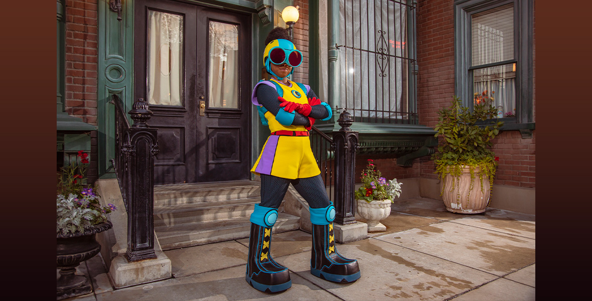  A promo image of Moon Girl from Marvel’s Moon Girl and Devil Dinosaur at Disney California Adventure Park. She’s standing in front of a brick building stoop and is wearing black and turquoise platform boots, a yellow shorts romper with purple accents, red gloves, turquoise goggles, and a yellow and turquoise helmet.