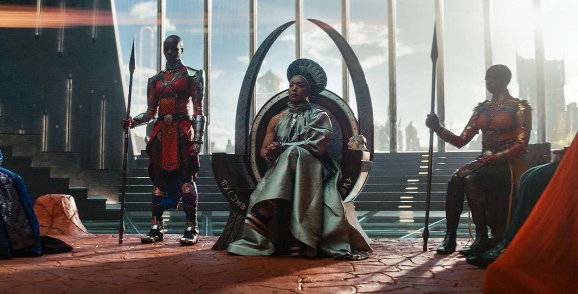 In a scene from Black Panther: Wakanda Forever, actor Angela Bassett as Queen Ramonda sits at the center in a large throne. She wears a silver dress and a headpiece. She is surrounded by two Dora Milaje warriors wearing red and gold armor and tunics.
