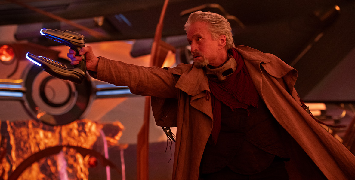 Hank Pym points a handheld futuristic weapon at something off-screen. He wears a brown coat with goggles around his neck. His white hair is brushed back.