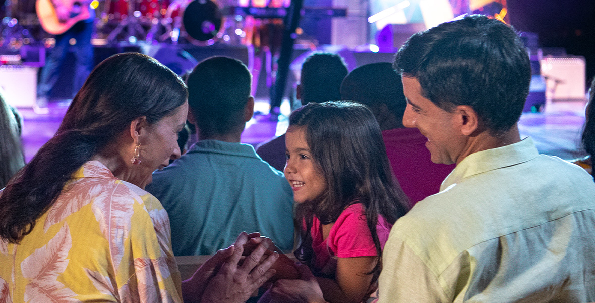 In a promotional image for the EPCOT International Flower & Garden Festival Garden Rocks Concert Series, a family of three is highlighted, enjoying a concert. The mother (left) has long dark hair and is wearing a yellow flowered shirt; she’s holding hands with her young daughter (center), also with long dark hair, wearing a pink shirt; the father (right) has short dark hair and is wearing a light yellow shirt. In the blurry background, a band is seen playing on a stage, and a few other audience members surround the family.