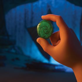 A view of Moana’s animated hand holding a small green stone between her thumb and pointer finger. The stone is embedded with the shape of a wave. In the background, a few ships are beached inside a dark cavern.