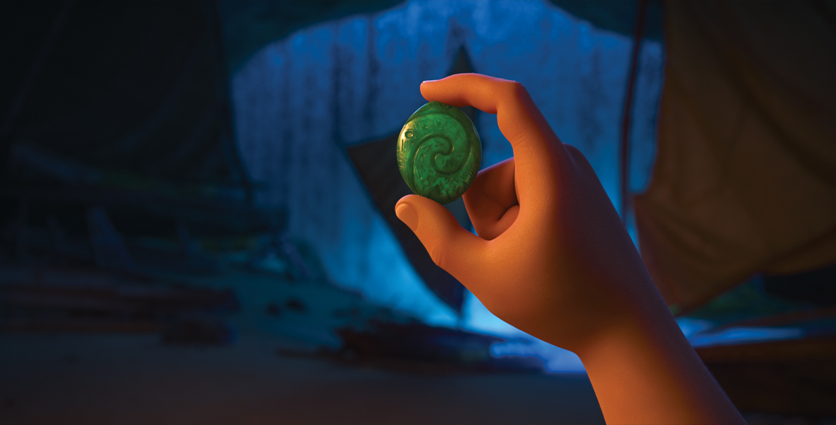 A view of Moana’s animated hand holding a small green stone between her thumb and pointer finger. The stone is embedded with the shape of a wave. In the background, a few ships are beached inside a dark cavern.