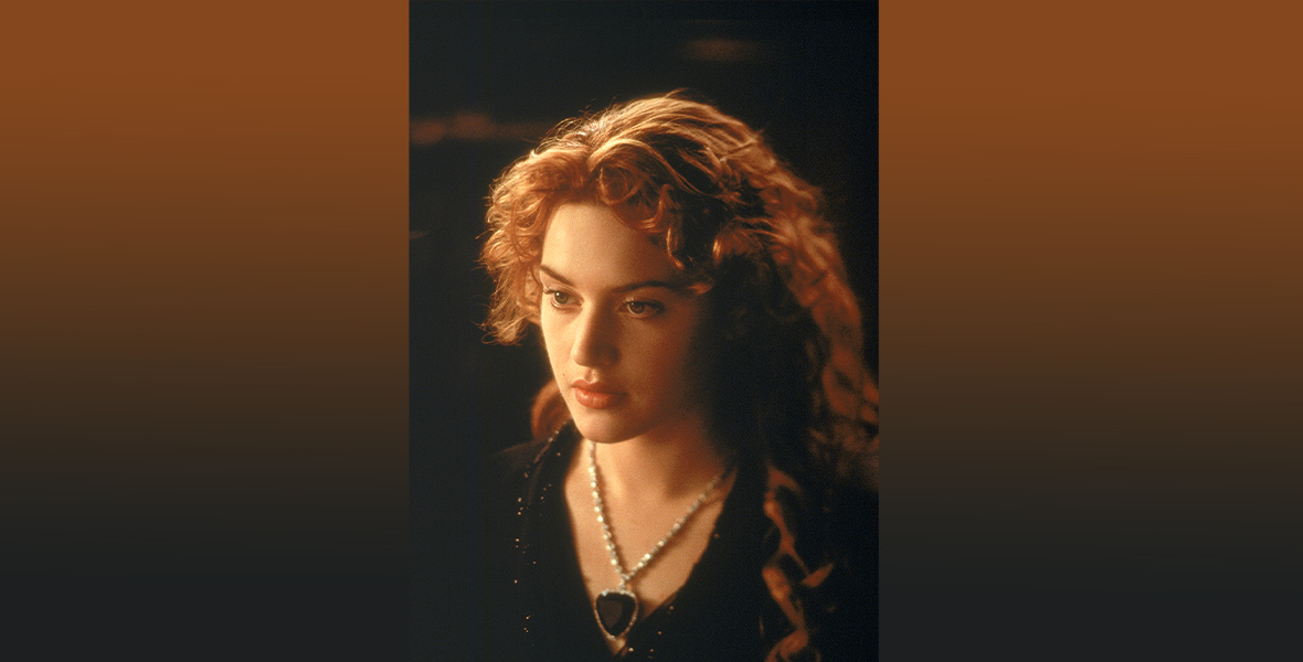 A close-up photo of Rose, played by Kate Winslet, wearing the Heart of the Ocean necklace. The heart-shaped, dark blue stone is attached to a silver chain around her neck. She looks slightly off-camera with a neutral expression. Her red hair is curled, half of it pulled back and away from her face.