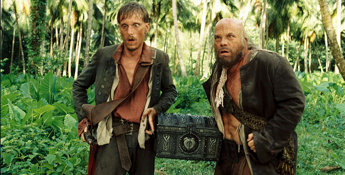 In a thick green jungle, two pirates look startled. The two men each have a hand on a midsize chest hoisted into the air between them. The chest looks to be made of iron with intricate markings on the top and sides. The pirates wear loose-fitting clothing and have their free hands on the swords at their sides.