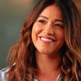 In a scene from Not Dead Yet, Gina Rodriguez wears blue and white striped pajamas. She has long, wavy brown hair and a big smile. A wine glass is in front of her.