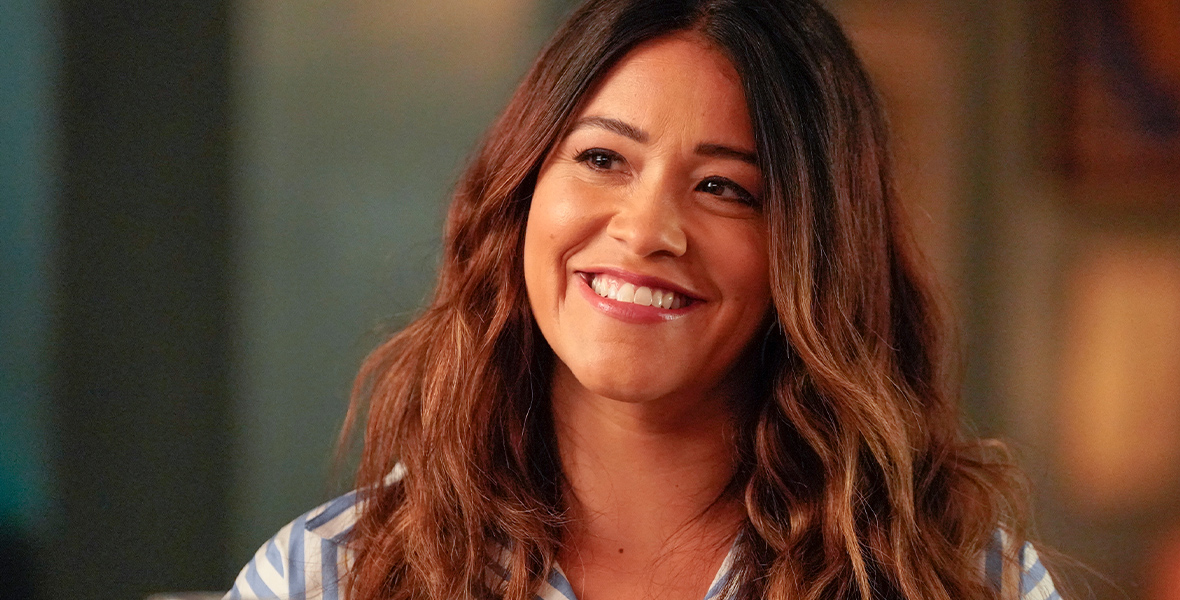 In a scene from Not Dead Yet, Gina Rodriguez wears blue and white striped pajamas. She has long, wavy brown hair and a big smile. A wine glass is in front of her.