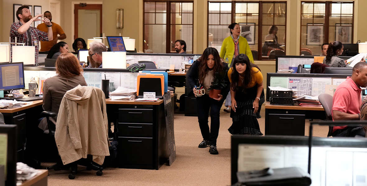In a scene from Not Dead Yet, Gina Rodriguez and Hannah Simone are hunched forward and walking through a bustling newsroom, surrounded by fellow writers, desks, computers, filing cabinets, and other common items found in an office.