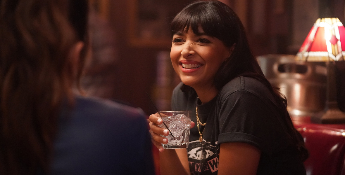 In a scene from Not Dead Yet, Hannah Simone wears a black T-shirt and a gold necklace. She is holding a glass in one hand and crinkling her nose as she smiles.