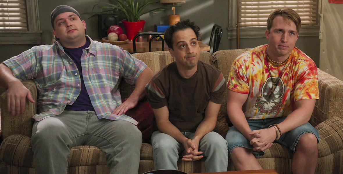 In a scene from an episode of The Goldbergs, three adult men sit on a brown plaid couch in an apartment.