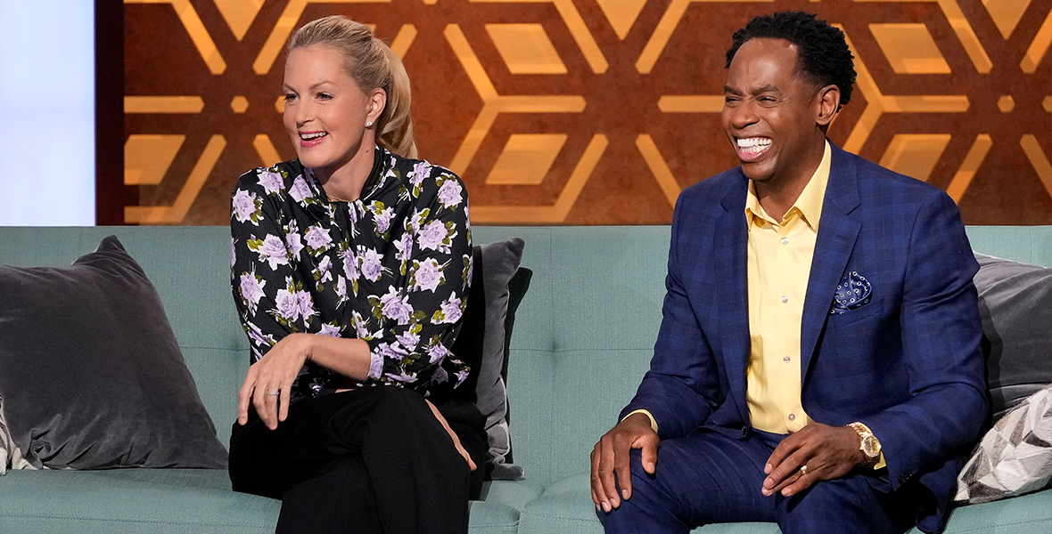 In a scene from an episode from The Parent Test, hosts Ali Wentworth and Dr. Adolph Brown III sit on a green couch. Wentworth wears black pants and a floral blouse with purple flowers. Brown wears a blue plaid suit and a yellow button-down shirt.