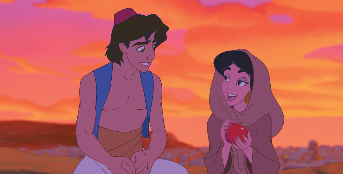 The animated Aladdin and Jasmine sit and talk on the edge of a wall, the orange and pink sky behind them. Jasmine wears a brown cloak and wrap, holding a red apple between her hands. Aladdin looks down at her, a red fez atop his black hair.