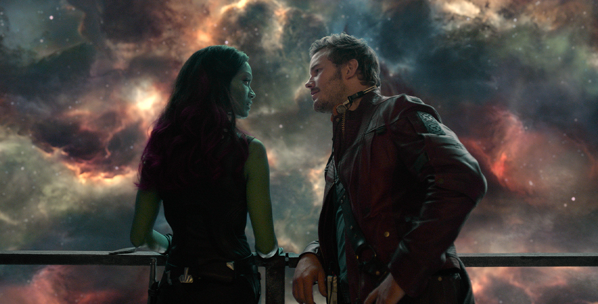 A shot of Peter and Gamora standing by a railing, a brilliant starry galaxy behind them. Gamora’s back is to the camera, but she looks to her right at Peter. Peter leans sideways against the railing, staring at Gamora with his headphones around his neck.