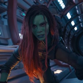 Gamora, a green-skinned assassin with purple hair, crouches down on the interior of a spaceship. She is wearing a skintight, utilitarian black jumpsuit and boots.