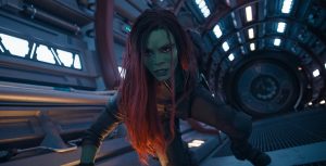 Gamora, a green-skinned assassin with purple hair, crouches down on the interior of a spaceship. She is wearing a skintight, utilitarian black jumpsuit and boots.