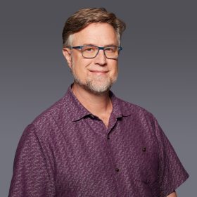 Dan Povenmire wears a purple, patterned polo shirt and blue eyeglasses. He is smiling and both of his hands are in his pockets as he poses against a gray backdrop.
