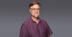 Dan Povenmire wears a purple, patterned polo shirt and blue eyeglasses. He is smiling and both of his hands are in his pockets as he poses against a gray backdrop.
