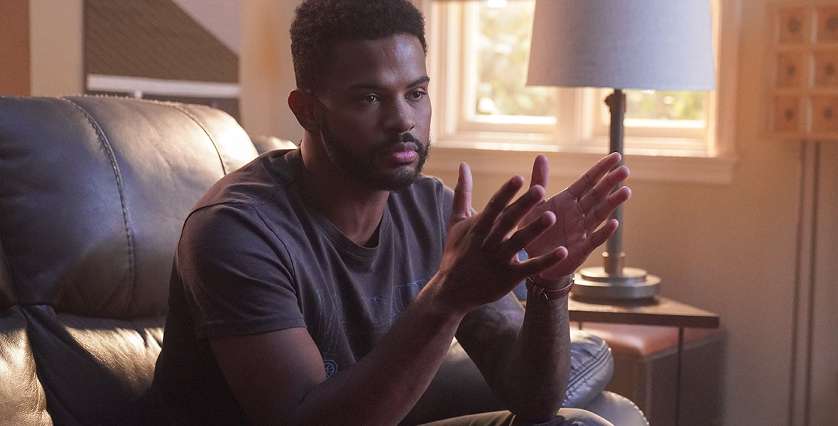 In a scene from an episode of Freeform’s grown-ish, actor Trevor Jackson as Aaron sits on a black leather couch and wears a gray T-shirt and denim jeans.