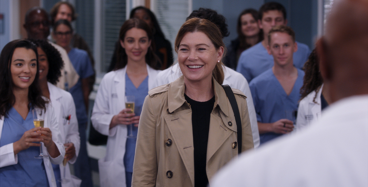 In a scene from an episode of ABC’s Grey’s Anatomy, actor and Disney Legend Ellen Pompeo as Meredith Grey stands center and is surrounded by doctors holding glasses of champagne. She wears a tan trench coat and a black T-shirt.