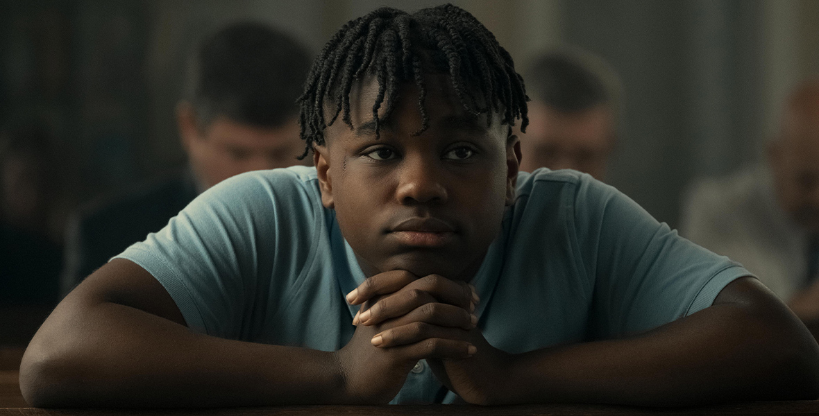 In the feature film Bruiser, actor Jalyn Hall as Darious leans on a wooden bench and wears a light blue T-shirt.